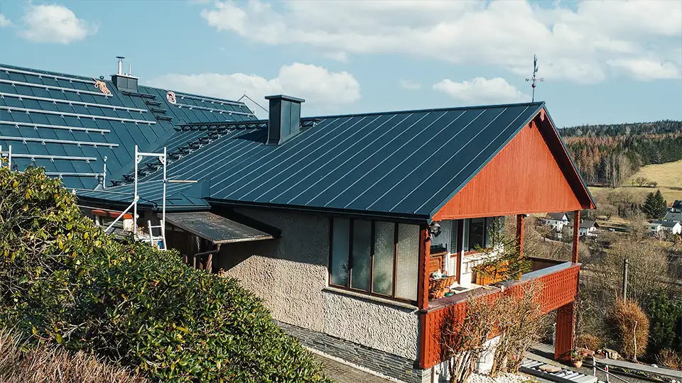 House with blue metal roof and red wooden balcony railing surrounded by trees and countryside.