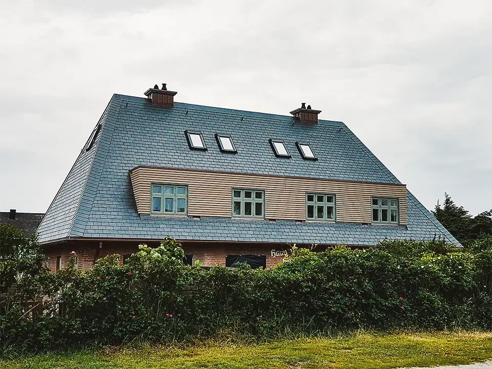 Large white modern house with multiple dormer windows surrounded by green bushes and trees.