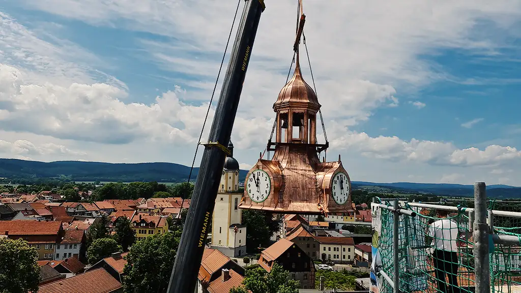 Crane lifting a large copper clock tower over a town with orange rooftops and green hills in the background.