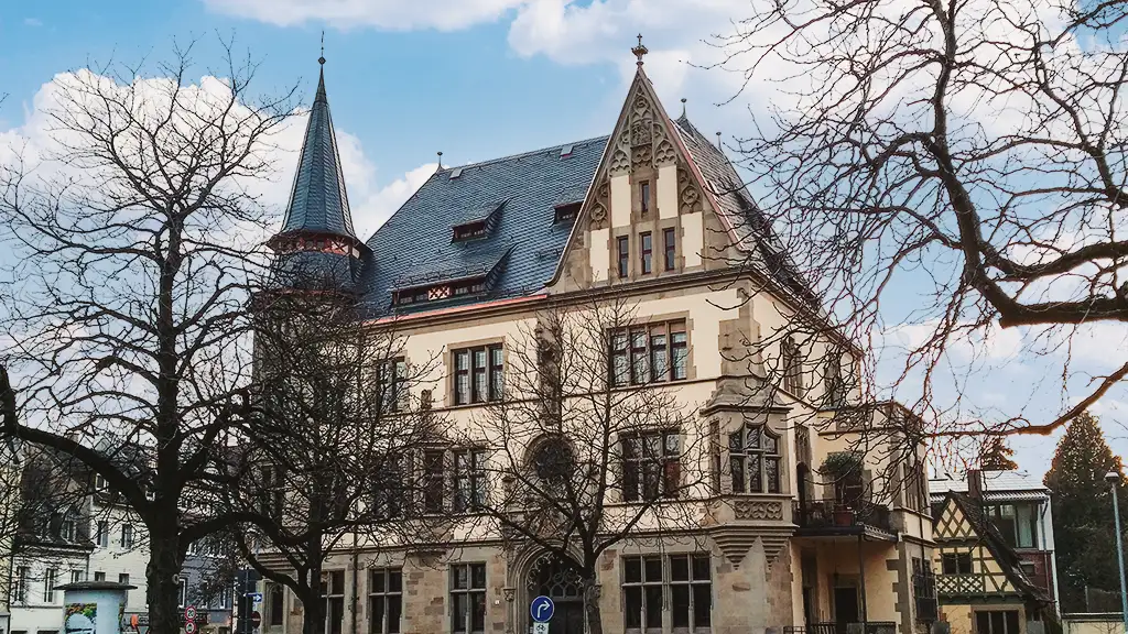 Historic multi-story building with steep slate roof and bare trees in front under a partly cloudy sky.