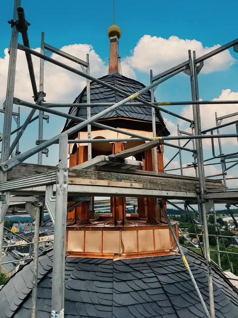 Rooftop tower with scaffolding, slate tiles, and a decorative finial against a blue sky with clouds.