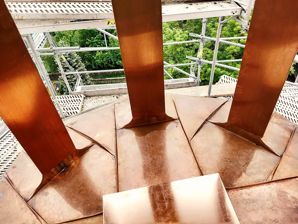 Copper roof structure with vertical supports viewed from above with scaffolding and trees in background.