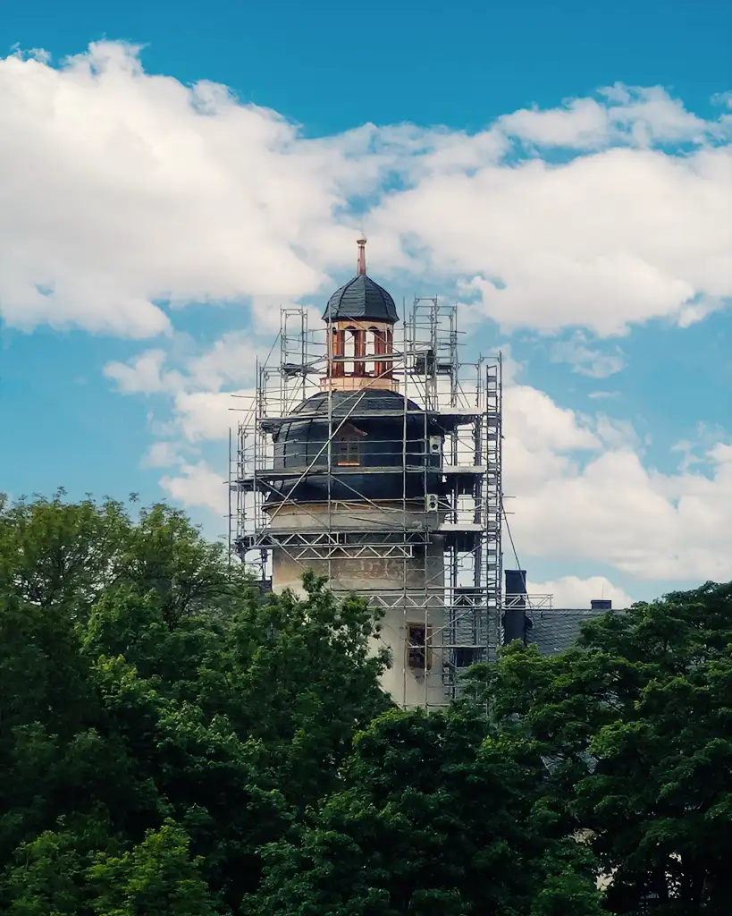 Historic tower surrounded by scaffolding above dense green trees under a blue sky with clouds.